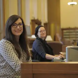Two women with brown hair smile from behind a desk in the library's reading room.