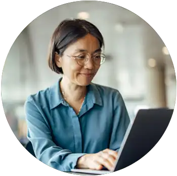 A middle-aged, medium-skin-toned Asian woman with glasses smiles and uses a laptop on a table.